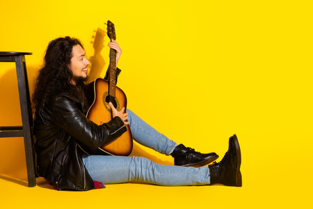 Young man with a guitar seated against a yellow background wearing a leather jacket and boots,...