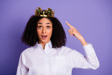 Confident young woman in a stylish white shirt wearing a crown pointing upward on a violet backgroundの写真素材
