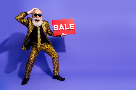 Chic elder man in gold-patterned suit holding a sign on vibrant purple background showcasing style and positivityの写真素材