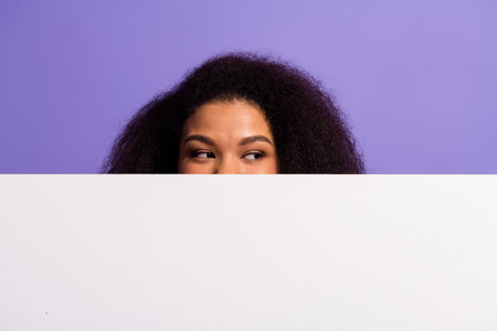 Young woman with curly hair peeking over a blank sign against a purple background, with a charming...