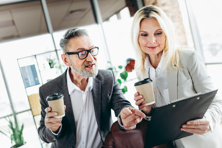 Senior professionals reviewing documents while enjoying coffee in a modern office setting emphasizing teamworkの写真素材