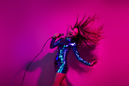 Joyful woman singing in a shiny blue dress with radiant blonde hair against a vibrant pink...