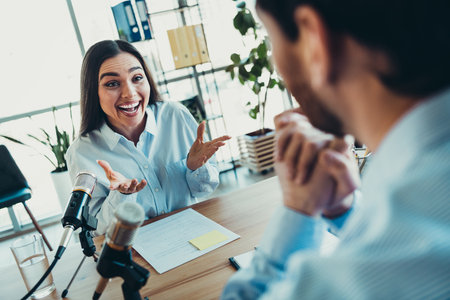 Young professional woman conducting an engaging podcast interview with a coworker in a workspace settingの写真素材