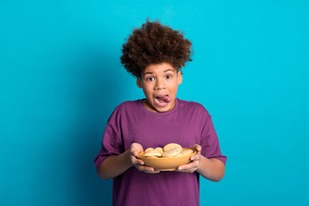 Cheerful boy holding a bowl of cookies against a vibrant blue background expressing a playful moodの写真素材