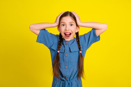 Excited young girl in a blue outfit holding her head, expressing joy on a vibrant yellow backgroundの写真素材