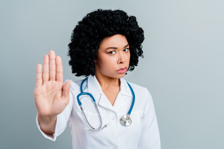 Skilled female doctor in white coat showing a stop gesture with her hand against light grey backgroundの写真素材
