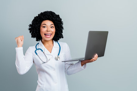 Skilled female doctor holding laptop celebrating success in healthcare on grey backgroundの写真素材