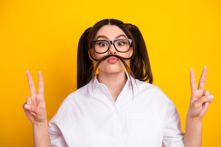 Playful young woman in white shirt making funny face and peace signs against a yellow background demonstrating a cheerful moodの写真素材