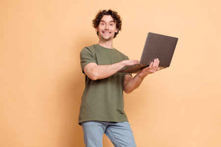 Young male with curly hair in khaki t-shirt using laptop against beige backdrop showcasing casual and stylish characterの写真素材
