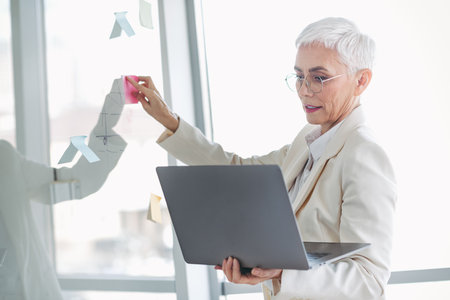 Successful mature businesswoman working with laptop and planning ideas using sticky notes in a bright modern officeの写真素材