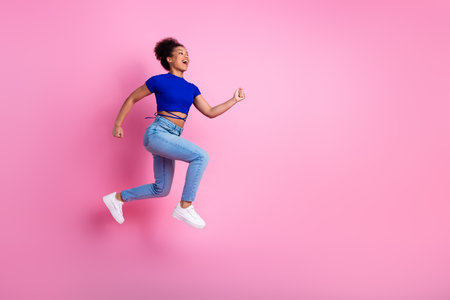 Happy young woman in trendy blue top and jeans enjoying an energetic jump against a pastel pink...