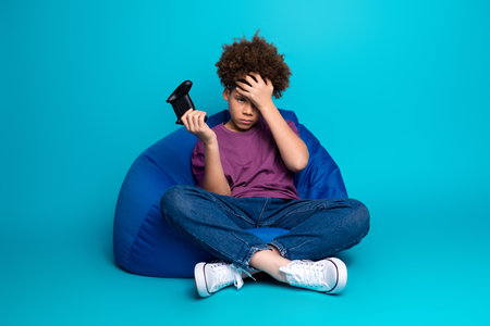 Young boy sits on a bean bag chair holding a game controller, appearing thoughtful against a vibrant...
