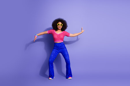 Energetic young woman in vibrant retro clothing dancing against a purple background, showcasing...