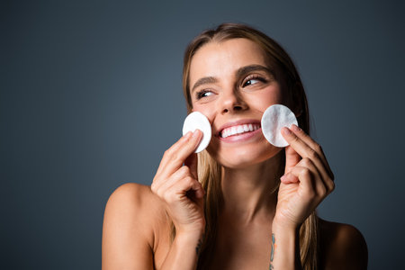 Young woman with radiant smile holding cotton pads exemplifying skincare and selfcare on a neutral backgroundの写真素材