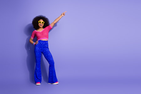Stylish young woman with vibrant fashion and afro hairstyle posing joyfully on a purple background,...