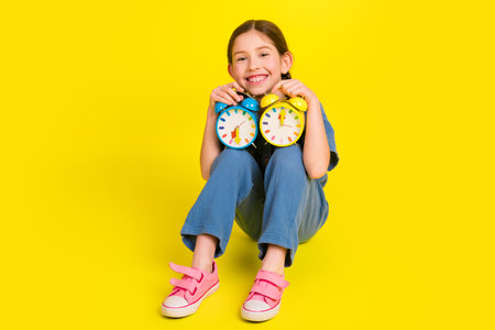 Smiling young girl holding colorful alarm clocks against vibrant yellow backdrop, showcasing...