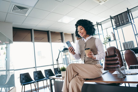 Young businesswoman in professional attire working in a stylish office environment while using her smartphone and enjoying coffee.の写真素材