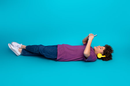 Young boy enjoying music and mobile gaming against a vibrant blue background while wearing yellow headphones and casual clothesの写真素材