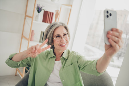 Beautiful elderly woman with gray hair taking a cheerful selfie gesture in a cozy home interior