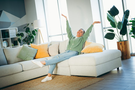 Young woman relaxing happily on a cozy sofa in a modern apartment surrounded by plants and bright natural daylightの写真素材
