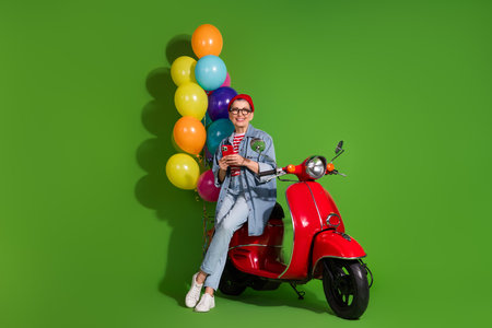 Fashionable senior woman relaxing on a red scooter, holding a smartphone, surrounded by colorful balloons in a vibrant green studio.の写真素材