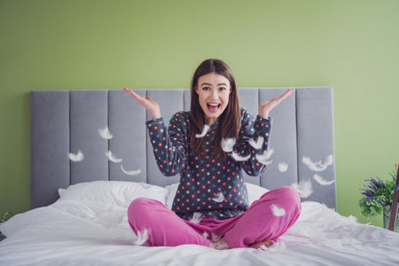 Young woman in playful mood tossing feathers while sitting on a bed in cozy pajamas amidst a cheerful and bright bedroom settingの写真素材