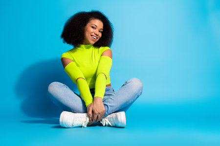 Stylish young woman sitting against a bright blue background wearing neon green top and denim jeans