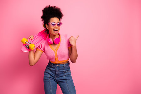 Stylish young woman posing with skateboard and headphones against vibrant pink background,...