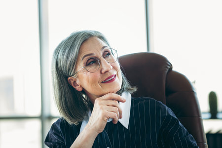 Confident mature businesswoman with gray hair in office chair wearing formal attire and glasses, exuding eleganceの写真素材