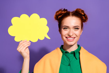 Smiling young woman with red hair buns holding a speech bubble against a vibrant purple backgroundの写真素材
