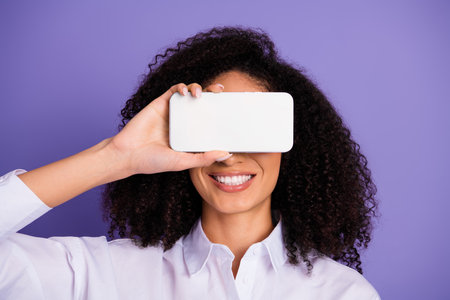 Smiling young woman holding a smartphone over her eye against a purple background, showcasing style and technologyの写真素材