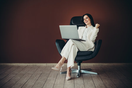 Beautiful businesswoman in white formalwear working on laptop while seated in a modern chair in a professional settingの写真素材