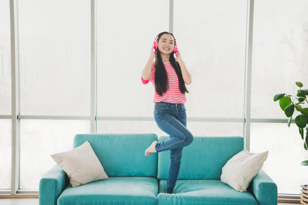 Young woman enjoying music with headphones on a sofa in a modern sunny interior, expressing happiness and relaxationの写真素材