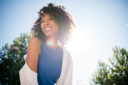 Charming young woman smiling brightly under the sunny sky enjoying urban outdoor leisure in stylish fashionの写真素材