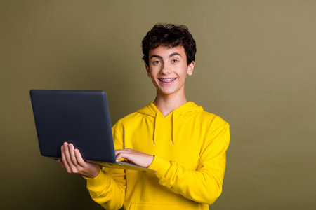 Smiling teenage boy wearing yellow hoodie holding laptop against khaki backgroundの写真素材