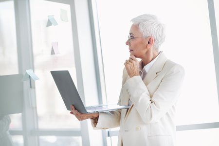 Confident senior businesswoman in elegant white suit analyzing strategy while using laptop in bright modern officeの写真素材