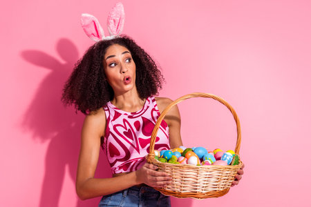 Young woman holding an Easter basket filled with colorful eggs, wearing festive bunny ears and a...