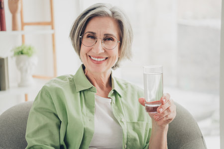 Portrait of a smiling mature woman with glasses, holding a glass of water while relaxing comfortably at homeの写真素材