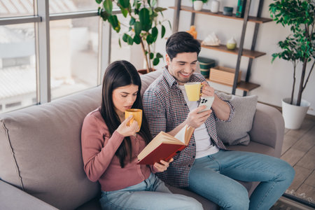 Young couple relaxing at home, enjoying leisure time together by reading and browsing in a cozy living room setting during the daylightの写真素材