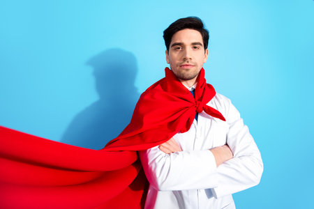 Confident male doctor in red cape and medical coat standing with arms crossed against light blue background portraying heroismの写真素材