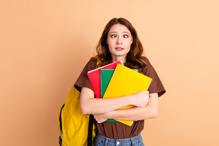 Confused young woman holding colorful notebooks against beige background, wearing casual brown shirt...