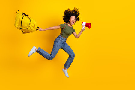 Energetic young woman jumping with a backpack and megaphone on a vibrant yellow background for promotionの写真素材