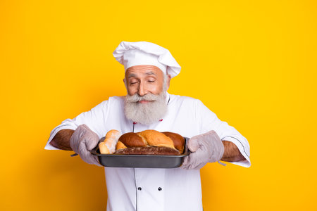 Elderly chef presenting freshly baked bread in front of a vibrant yellow backgroundの写真素材