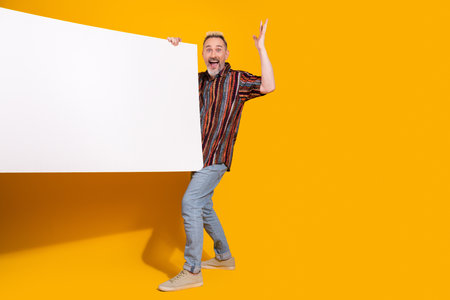 Smiling middle aged man holding a white display board against a vibrant yellow background, dressed...
