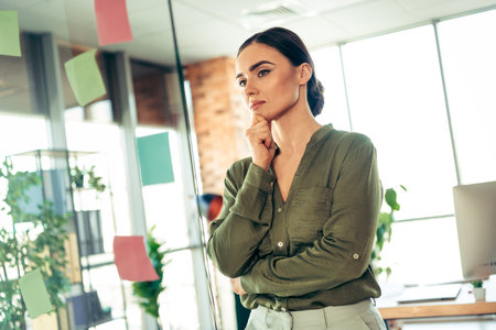 Thoughtful businesswoman in a modern office reflecting on ideas surrounded by glass walls and colorful notesの写真素材