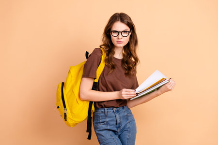 Young woman with long brown hair wearing casual clothing and yellow backpack, holding notebooks against a beige backgroundの写真素材