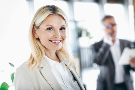 Smiling businesswoman posing in a modern office environment with a businessman coworker in the background making a callの写真素材