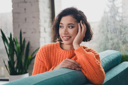 Portrait of a young woman in a bright orange sweater enjoying a relaxing moment indoors on a cozy dayの写真素材