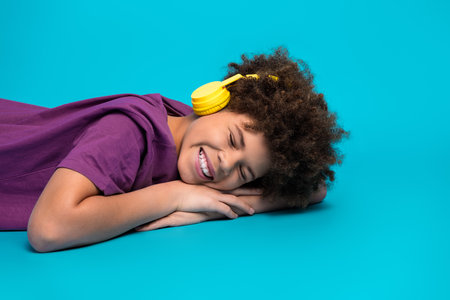 Smiling young boy with curly hair relaxing and listening to music on yellow headphones against a turquoise backgroundの写真素材