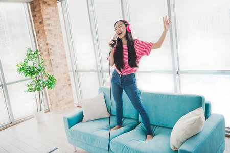 Young woman enjoying a karaoke session at home, standing joyfully on the couch while singing with a microphone and headphonesの写真素材